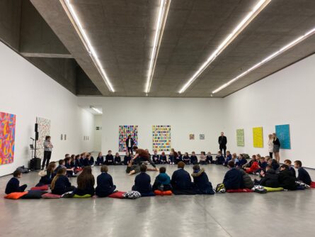 a group of children sitting on cushions in a square. In the centre of the square, two people are performing a dance work. The walls are hung with paintings.